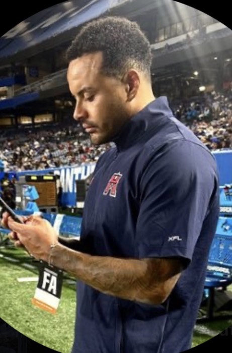 Hiram Velez on a sideline in team staff gear, working from his phone.